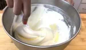 Mixing whipped cream and butter in a metal bowl with a spatula, close-up on hand and ingredients.